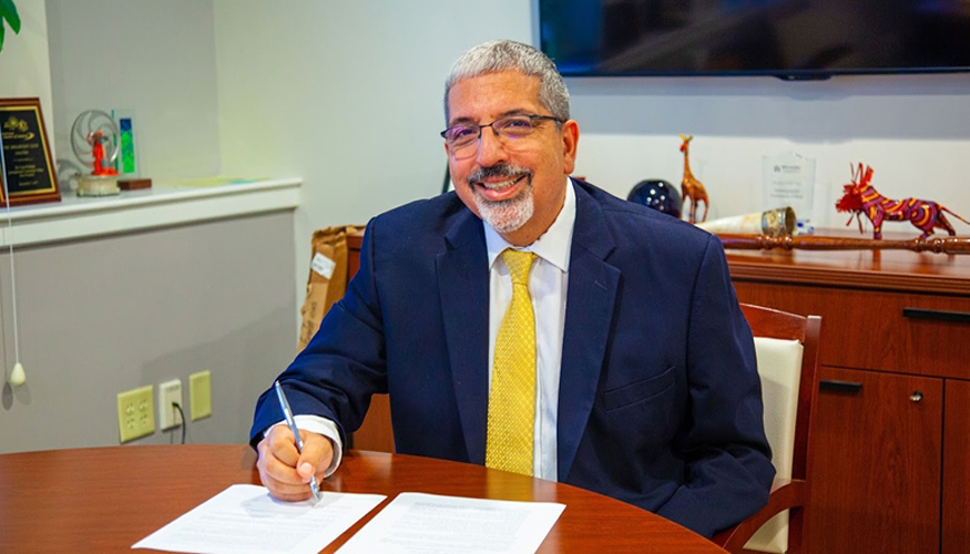 Quinsigamond Community College President Luis Pedraja at his desk smiling with papers