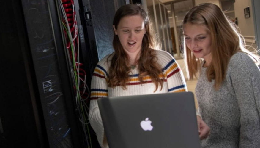 two students standing at laptop screen together, with cybersecurity infrastructure in background